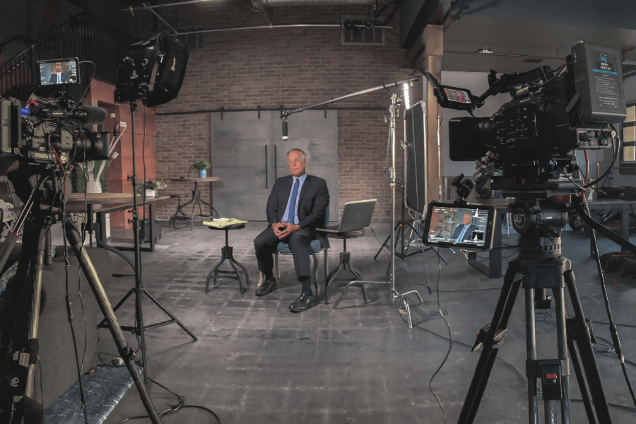 a man in a suit is sitting down, with cameras and video equipment pointed at him