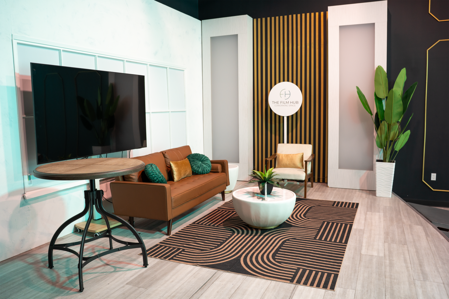 LED-lit modern stage set at The Film Hub featuring a tan sofa, accent pillows, coffee table, lounge chair, large TV monitor, and decorative backdrop with vertical slats and branding.