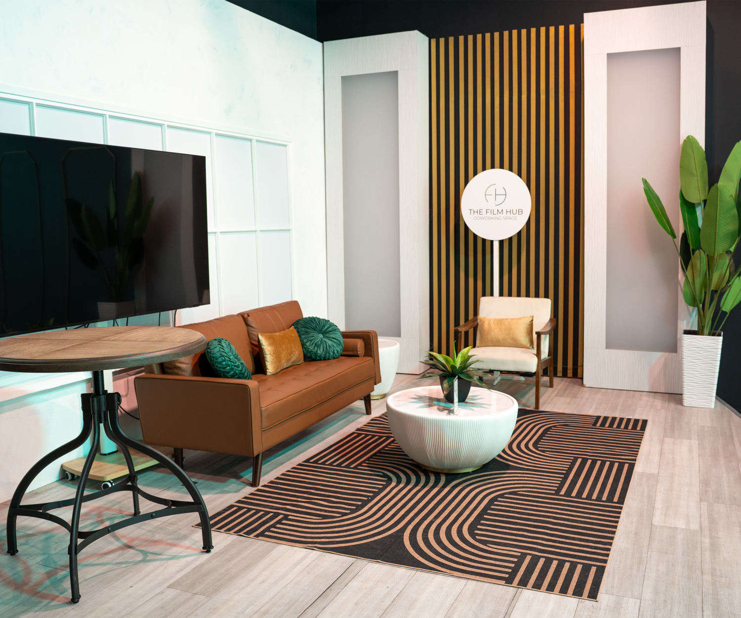 LED-lit modern stage set at The Film Hub featuring a tan sofa, accent pillows, coffee table, lounge chair, large TV monitor, and decorative backdrop with vertical slats and branding.