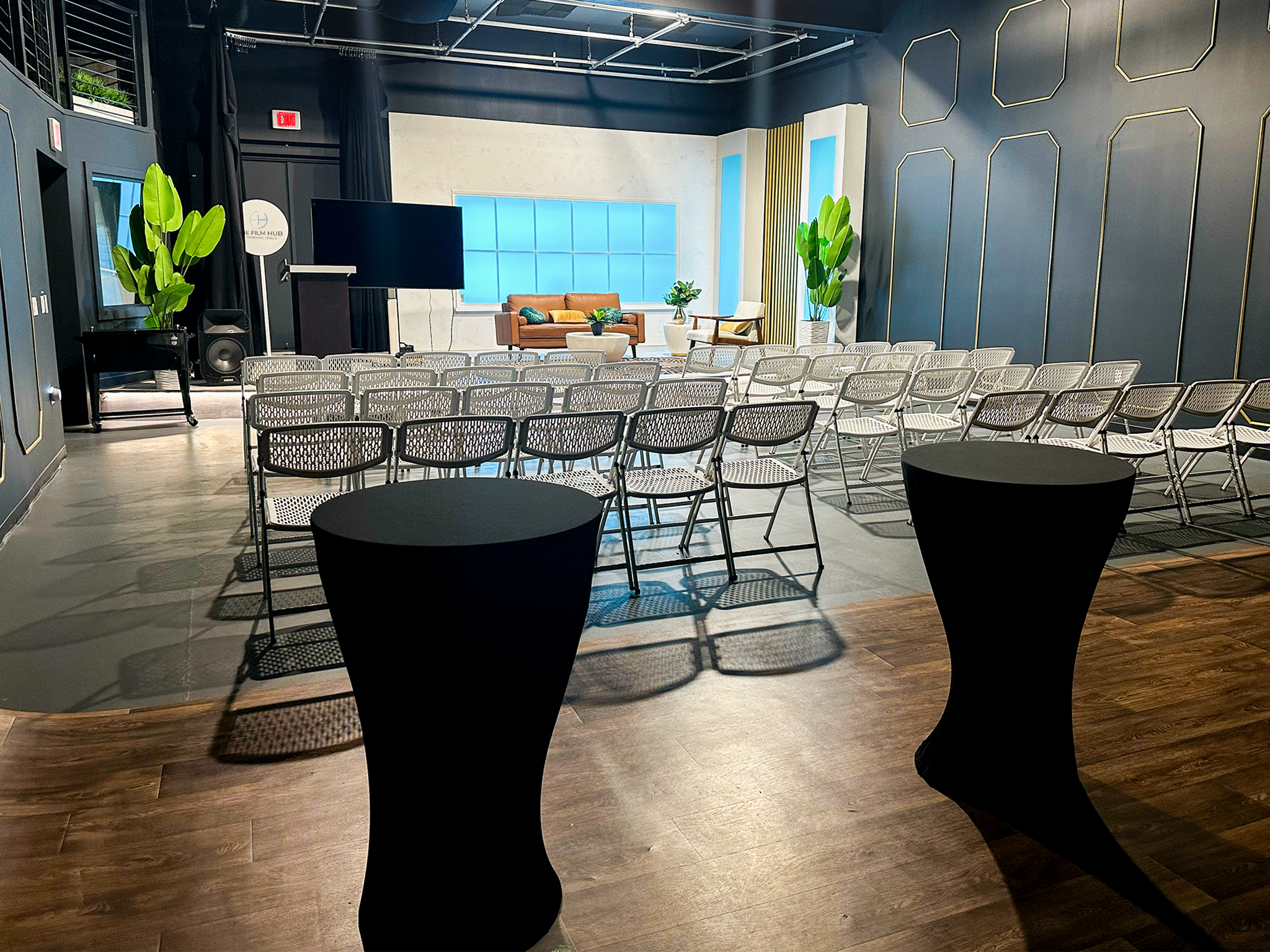 Stage 2 event setup at The Film Hub with rows of white chairs facing a podium, LED-lit backdrop in blue, modern lounge seating, and cocktail tables in the foreground.
