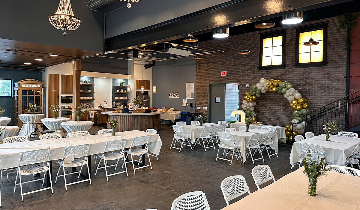 Event setup in Audrey Hepburn Hall at The Film Hub, featuring banquet tables with white linens, floral centerpieces, cocktail tables, a full kitchen, and balloon arch decor in a modern industrial-style venue.