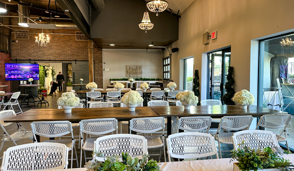 Chic event setup in Audrey Hepburn Hall at The Film Hub, featuring long wooden tables, white chairs, and elegant white floral centerpieces under crystal chandeliers with natural light from large windows.